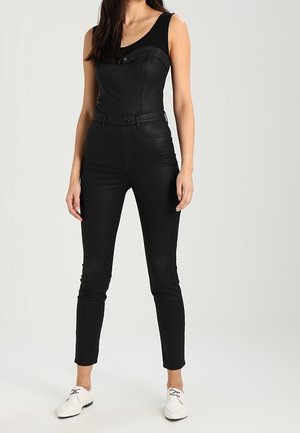 Kvinde iført sort ærmeløs jumpsuit med knapdetalje og hvide snøresko mod ensfarvet baggrund.