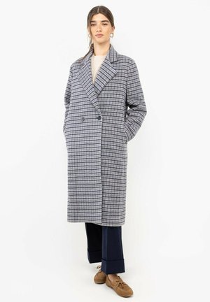 Mujer con un abrigo largo gris a cuadros, jersey beige, pantalones azul marino remangados y mocasines marrones con cordones, de pie frente a un fondo blanco.