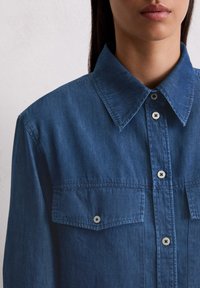 Blauwe spijkerbloes met een spitse kraag, sluiting met knopen aan de voorkant en twee borstzakken. Glad materiaal en contrasterende witte knopen.