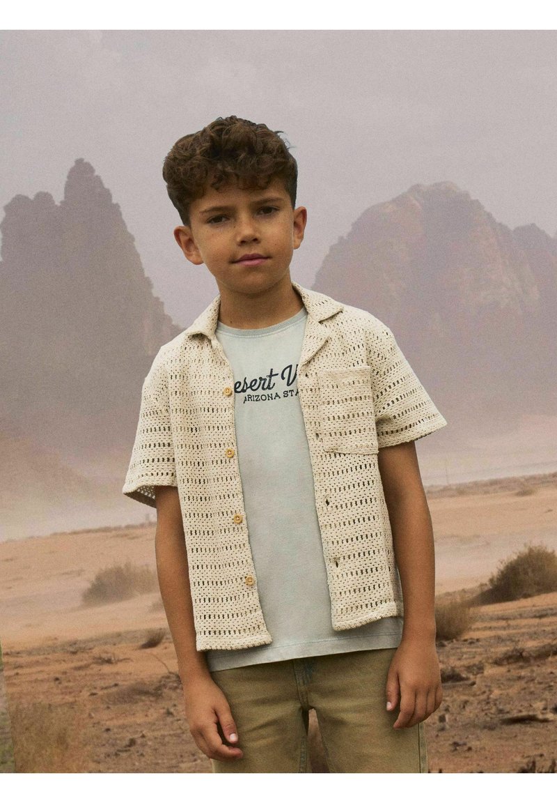 Chemise en tricot à manches courtes couleur crème avec un motif texturé et des boutons en bois, portée sur un t-shirt graphique bleu clair, associée à un pantalon beige.