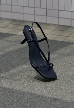 Schwarze, hochhackige Sandale mit dünnen Riemchen und einer goldenen Schnalle, die aufrecht auf einem strukturierten Gehweg nahe einer gefliesten Wand steht.