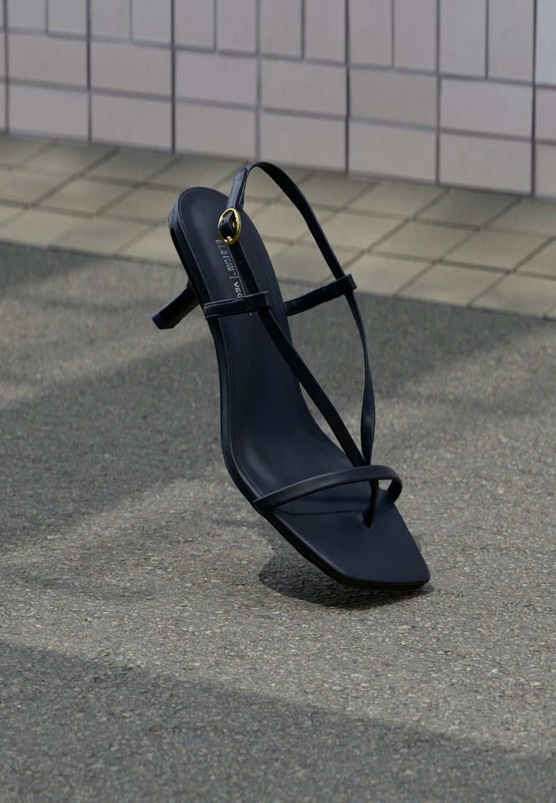 Μαύρο ψηλοτάκουνο σανδάλι με λεπτούς ιμάντες και χρυσή αγκράφα, όρθιο σε υφή πεζοδρομίου κοντά σε πλακιδωτό τοίχο.