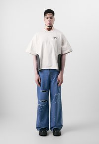 Beige Cropped-Shirt mit grauen Seiteneinsätzen; lässige blaue Jeans mit ausgefransten Rissen und Farbspritzern; schwarze klobige Schuhe.