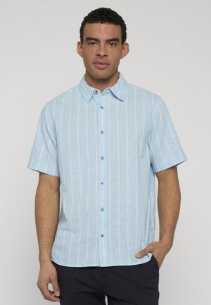 Homme portant une chemise à manches courtes bleu clair à fines rayures blanches verticales, debout avec une main dans la poche.