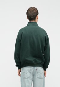 Tmavě zelená fleecová mikina s vysokým límcem, oversized střihem a žebrovanými manžetami, kombinované se světle modrými džíny s kapsami na zadní straně.