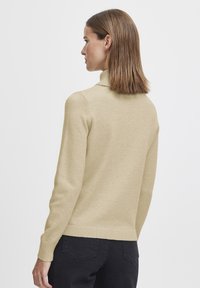 Beige polotröja med långa ärmar, ribbade �ärmslut och fåll. Mjuk stickad textur. Buren med svarta byxor, sedd från ryggen.