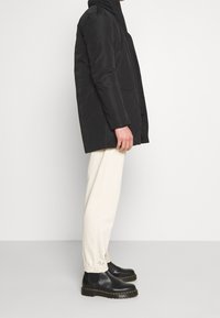 Manteau noir rembourré avec une coupe droite, poches latérales et col haut. Porté avec un pantalon crème et des bottines noires.