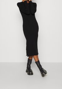 Robe tricot ribbed noire avec des manches longues, un col rond et des fentes latérales. Associée à des bottines noires à semelle épaisse.