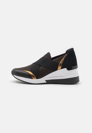 Schwarzer Slip-On-Sneaker mit goldenen Akzenten, weißer Zwischensohle, elastischen Kreuzriemen und Keilabsatz vor weißem Hintergrund.