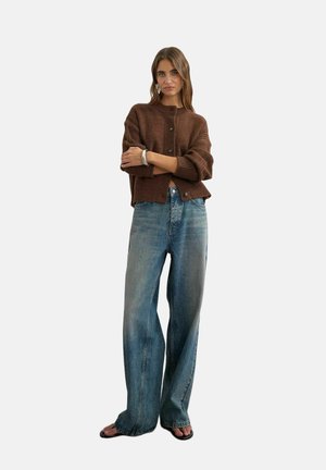 Femme portant un cardigan marron boutonné, un jean bleu délavé à jambes larges et des sandales noires, debout les bras croisés.