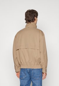 Chaqueta ligera color beige con un alto cuello, cinturilla elástica y solapas traseras, mostrada desde la parte posterior sobre un fondo blanco liso.