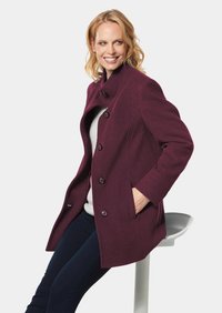 Bordeauxfarbener Wollmantel mit hohem Kragen, großen Knöpfen, Seitentaschen und einer strukturierten Form. Über einem leichten Pullover und dunklen Jeans getragen.