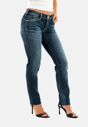 Jean en denim bleu foncé avec une coupe slim ; présente des accessoires en métal doré, des poches avant et une texture légèrement délavée. Associé à des talons hauts noirs.