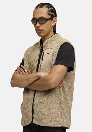 Mann mit geflochtenem Haar, der eine Sonnenbrille trägt und eine beige Puma-Fleeceweste über einem schwarzen T-Shirt trägt, die Arme verschränkt vor einem schlichten Hintergrund.