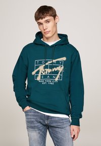 Sudadera de color verde oscuro con un bolsillo frontal, que presenta un logotipo gráfico beige que dice "TOMMY JEANS" y "NUEVA YORK EST. 1985."