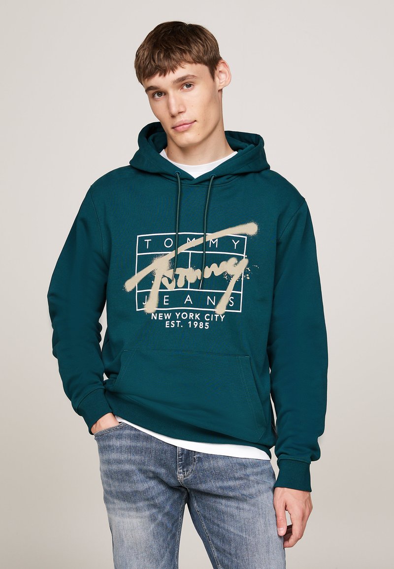 Sudadera de color verde oscuro con un bolsillo frontal, que presenta un logotipo gráfico beige que dice "TOMMY JEANS" y "NUEVA YORK EST. 1985."