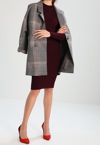 Manteau oversize à carreaux gris porté sur une robe côtelée bordeaux, accompagné de talons hauts rouges. Le manteau présente un col cranté et une coupe décontractée.