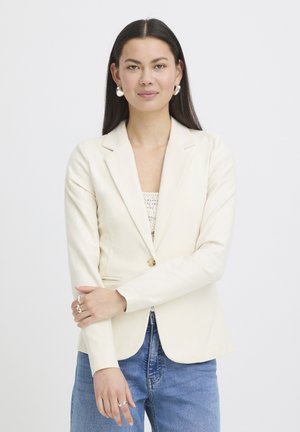 Kobieta o długich ciemnych włosach, ubrana w kremową marynarkę, biały koronkowy top, niebieskie dżinsy, srebrne kolczyki koła i srebrne pierścionki na białym tle.