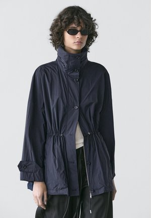 Krøllede person med sorte solbriller og en mørkeblå knappet jakke med indsnørret talje og høj krave, stående foran en ensfarvet baggrund.