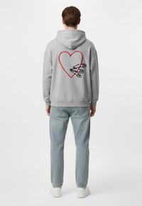 Felpa grigia con cappuccio e un grande design a forma di cuore rosso sul retro, caratterizzata da spille da balia nere. Indossata con jeans azzurrini e sneakers bianche.