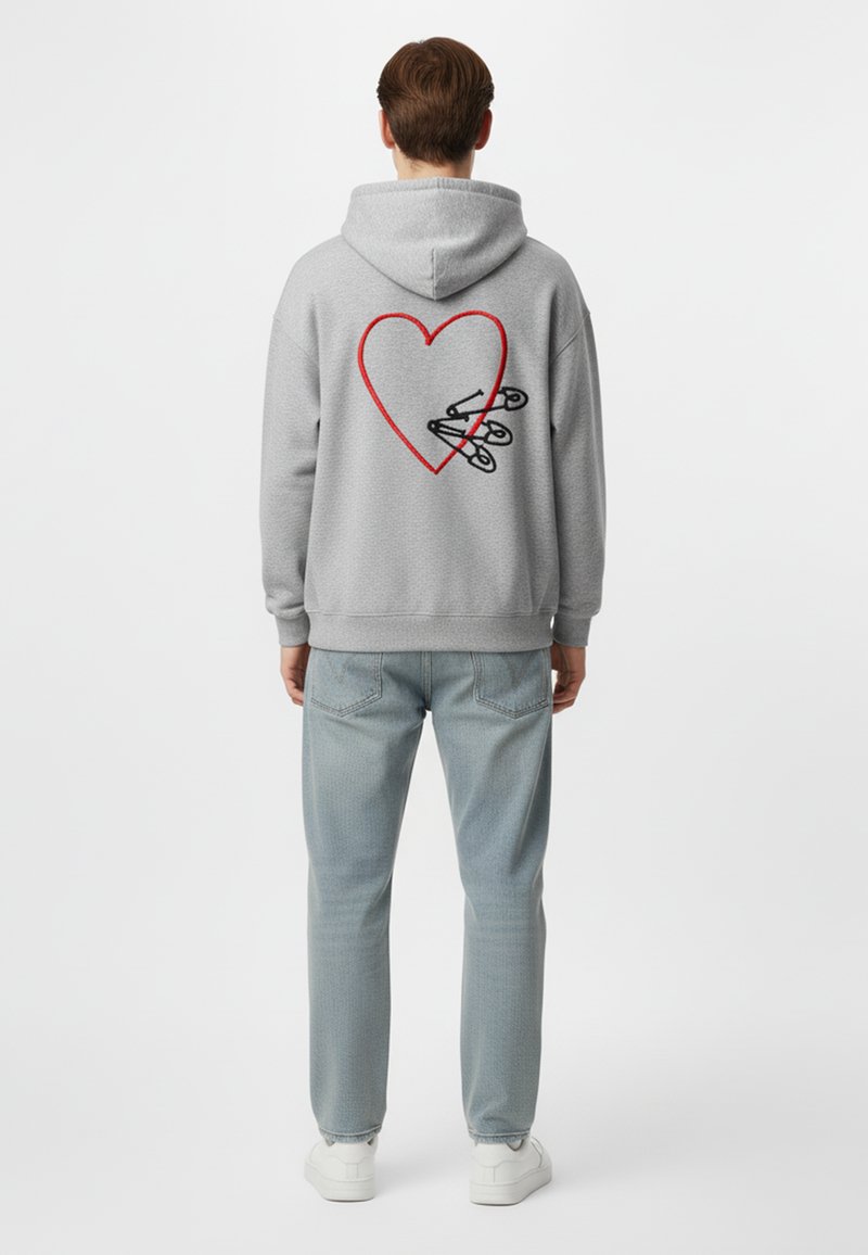 Felpa grigia con cappuccio e un grande design a forma di cuore rosso sul retro, caratterizzata da spille da balia nere. Indossata con jeans azzurrini e sneakers bianche.