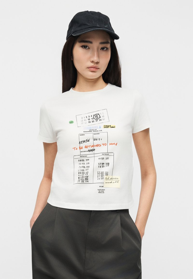 Jeune femme portant un t-shirt blanc court imprimé d'une carte de retour de livre de bibliothèque et une casquette noire, les mains dans les poches d'un pantalon sombre, sur fond blanc.