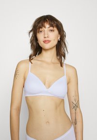 Sostén de color púrpura claro, sin costuras, con tirantes delgados ajustables, copas triangulares y una textura suave. Presenta un diseño simple sin herrajes ni patrones.