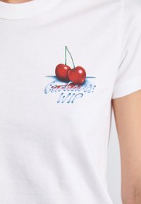 Camiseta de algodón blanca con un gráfico de dos cerezas rojas con tallos verdes y el texto "Carhartt WIP" en azul debajo. Mangas cortas.