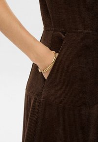 Rjava rebrasta obleka iz blaga z stranskimi žepi, s strukturno površino. Na zapestju so tri zlato obroči.