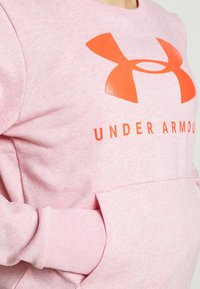 Růžová mikina s texturovaným povrchem, která má velké oranžové logo Under Armour a žebrované manžety. Obsahuje přední kapsu.