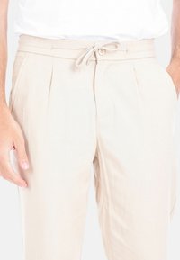 Pantaloni beige leggeri, plissettati, con vita elastica, chiusura con cordoncino e tasche laterali. Il tessuto testurizzato conferisce una finitura morbida.
