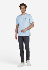Lichtblauwe katoenen t-shirt met een klein zwart palmbom ontwerp op de borst, gecombineerd met donkergrijze slim-fit broek en witte sneakers.