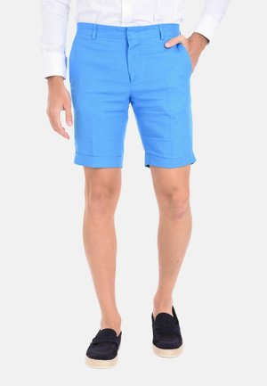 Uomo con pantaloncini blu brillanti con orlo arrotolato, camicia bianca e scarpe nere senza lacci, in piedi con una mano in tasca.