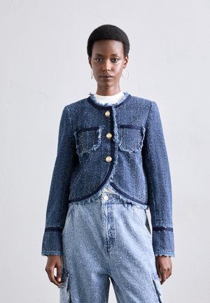 Denim-tweed takki, jossa on rispaantuneet reunat, laivastonsininen väri, pyöreä kaulus, kaksi etutaskua ja kultanapit. Yhdistetty vaaleansiinisiin teksturoituihin housuihin.