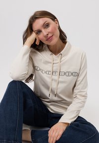 Beige hoodie met een kettingontwerp op de borst, voorzien van trekkoorden en lange mouwen, gecombineerd met donkere spijkerbroek.