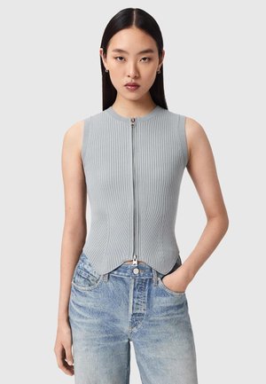 Nuori nainen, jolla on pitkät tummat hiukset, pukeutunut hihattomaan harmaaseen ribbineulottuun vetoketjutoppaan ja vaaleansinisiin farkkuihin, toinen käsi taskussa.