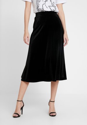 Vrouw met een zwarte fluwelen midi-rok, een wit T-shirt met print en zwarte hoge hakken met enkelbandje, staand tegen een effen achtergrond.