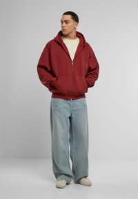 Maroon zip-up hoodie featuring a spacious hood, front pockets, and relaxed fit, paired with light blue wide-leg jeans and off-white sneakers.