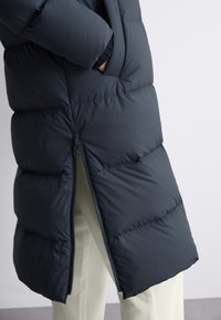 Mīksts tumši zils mētelis ar rāvējslēdzēja aizdari, stāvu apkakli un sānu kabatām. Materiāls ir gluds, ar spīdīgu virsmu un quilted dizainu.