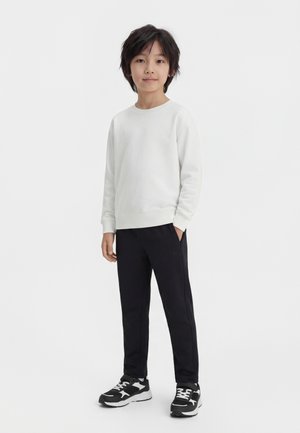 Ragazzo in piedi che indossa una maglia bianca a maniche lunghe, pantaloni neri e scarpe da ginnastica bianche e nere su uno sfondo chiaro uniforme.