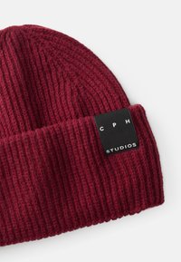 Beanie a maglia bordeaux con texture a coste e etichetta nera con la scritta "CPH STUDIOS" in bianco. Materiale morbido e caldo.