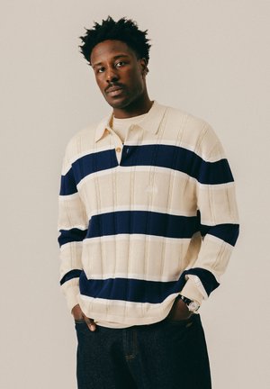 Homme aux cheveux courts et bouclés portant un pull crème à rayures bleu marine et un jean foncé, debout les mains dans les poches devant un fond uni.