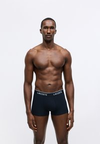 Black cotton boxer shorts feature a white "LACOSTE" logo on the elastic waistband. Model stands against a plain white background.