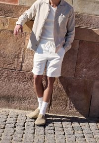 Chemise beige clair boutonnée portée sur un t-shirt blanc, associée à un short blanc et des chaussures grises. La tenue présente une coupe décontractée et un style casual.