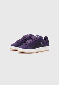 Baskets en suede violet avec des accents noirs et trois rayures noires, dotées de lacets plats et d'une semelle en caoutchouc blanche. Texture douce, forme classique.