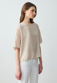 Beige top met korte mouwen en ronde hals, gemaakt van lichtgewicht materiaal, heeft een rechte snit en subtiele glans. Gecombineerd met witte broek.
