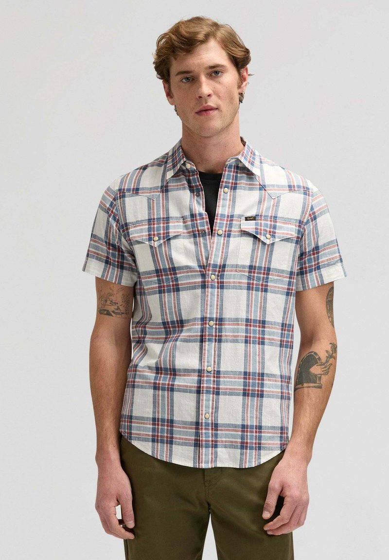 Jeune homme aux cheveux châtain clair portant une chemise à manches courtes à carreaux blanche, bleue et rouge, avec un pantalon vert olive, debout devant un fond uni.