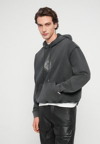 Νεαρός με καστανά μαλλιά, φορώντας ξεθωριασμένο μαύρο φούτερ με κουκούλα και μαύρο δερμάτινο παντελόνι cargo, στέκεται μπροστά σε απλό ανοιχτόχρωμο φόντο.