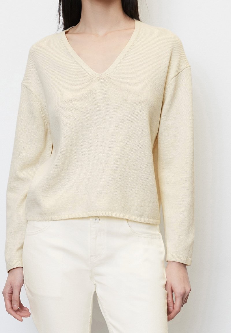 Femme portant un pull en maille crème à col en V et un pantalon blanc, debout devant un fond clair uni.