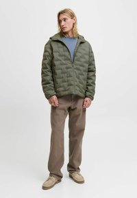 Mladá osoba v olivově zelené prošívané bundě, modré košili, volných hnědých kalhotách a béžových botách stojí na jednoduchém světlém pozadí.